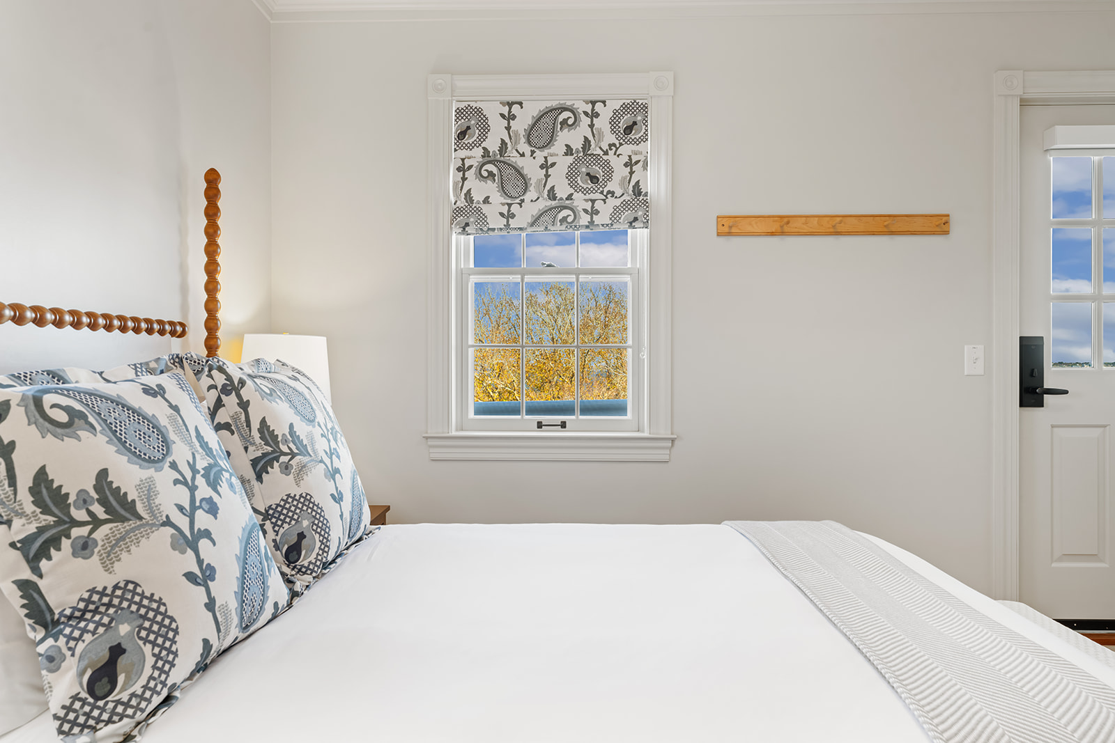 A wide view of a guest room with a white bed, blue and white pillows, a wooden headboard, and a window with a view of trees outside.