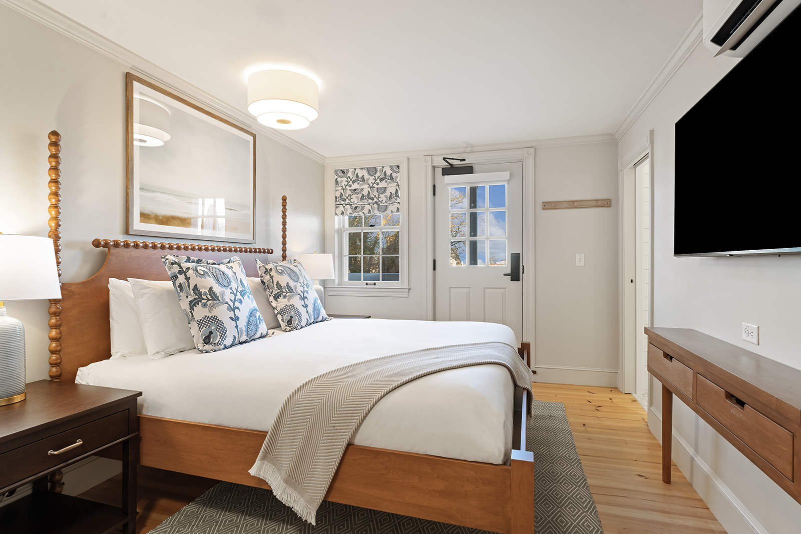 A wide view of a hotel room with a bed, nightstand, lamp, TV, window, and door.