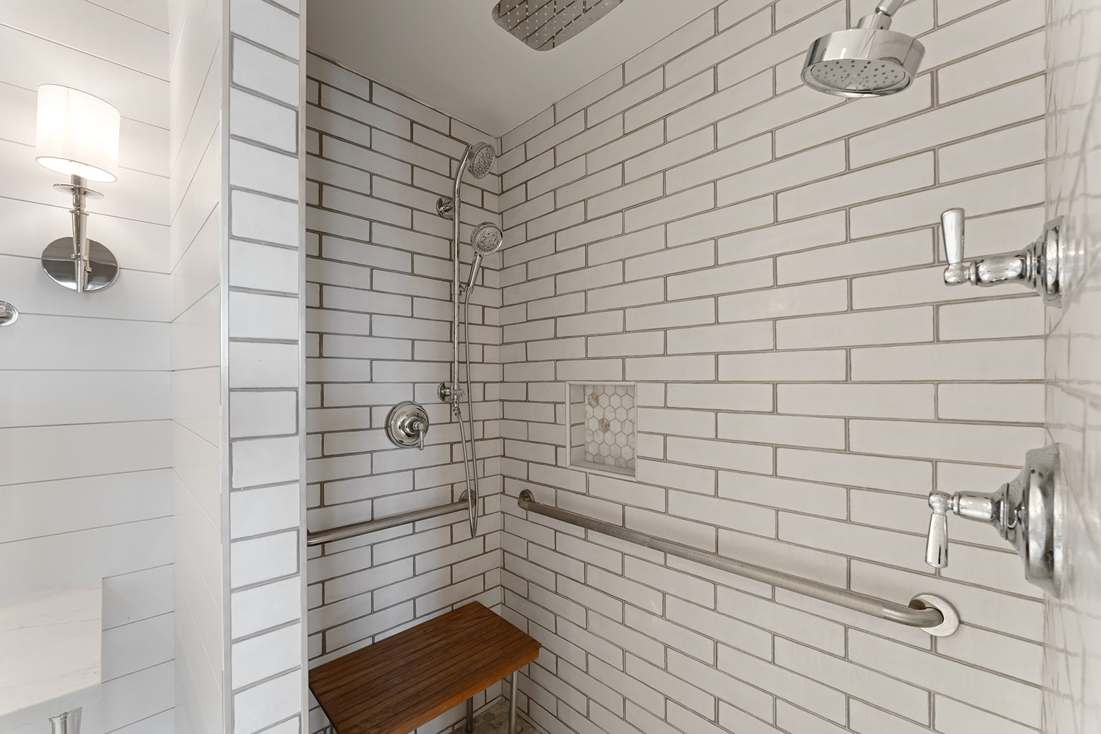 A clean and modern bathroom shower with white subway tiles, a wooden bench, and silver fixtures.