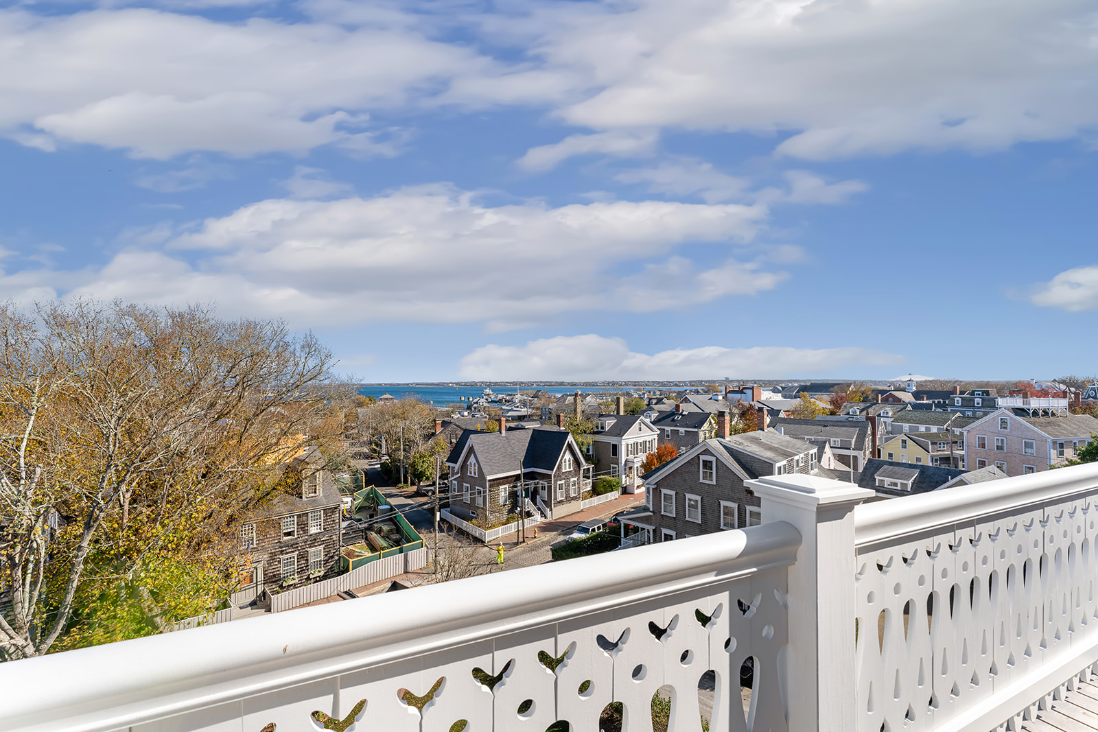 A white balcony railing overlooks a town with a body of water in the distance under a blue sky with clouds.