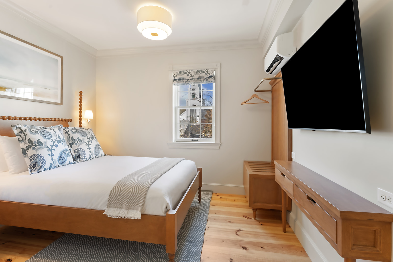 A neatly made bed with white and blue bedding in a light wood-floored room with a large flat-screen TV and a window