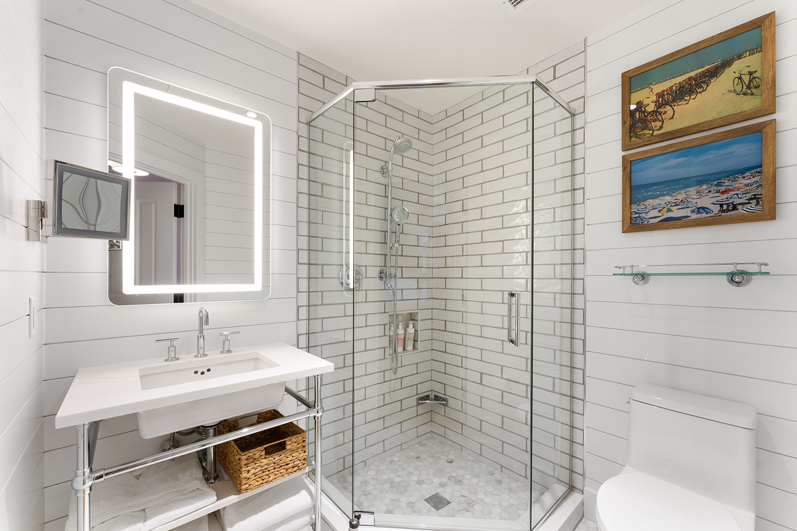 A modern bathroom with a glass-enclosed shower, double vanity, and toilet, featuring white tiles and a large illuminated mirror.